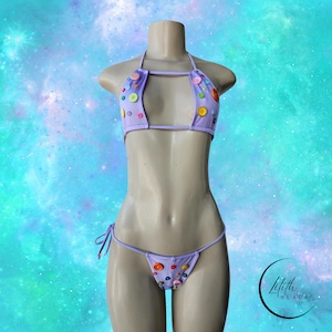 May include: A lavender bikini set with colorful buttons. The halter top and string bikini bottom are adorned with various sized buttons in shades of yellow, pink, green, and blue. The bikini is displayed on a mannequin against a blue and green background.
