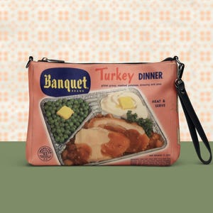 Puede incluir: Un bolso de mano rosa con un diseño retro de "Banquet Turkey Dinner". La imagen muestra una cena televisada con pavo, puré de patatas, guisantes y salsa. Tiene una correa de muñeca negra y un cierre de cremallera.