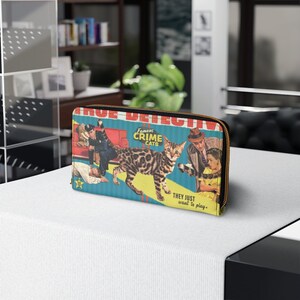 Pu&ograve; includere: Un portafoglio con cerniera blu e giallo con un'illustrazione vintage di un gatto e persone. Il testo sul portafoglio recita "Famous Crime Cats" e "They just want to play."