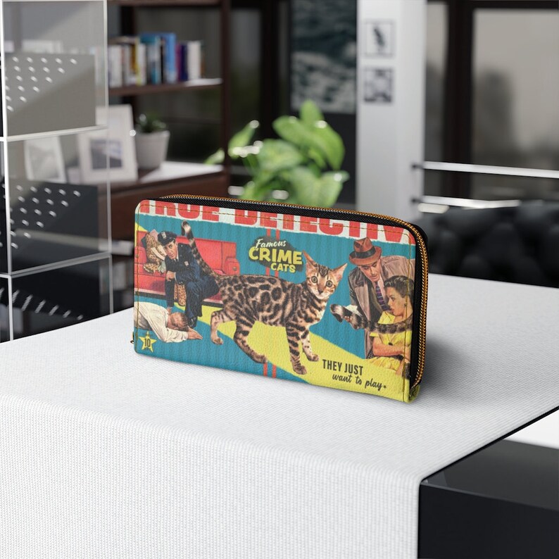 Pu&ograve; includere: Un portafoglio con cerniera blu e giallo con un'illustrazione vintage di un gatto e persone. Il testo sul portafoglio recita "Famous Crime Cats" e "They just want to play."