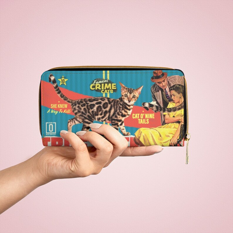 Pu&ograve; includere: Un portafoglio con cerniera blu e rosso con un'illustrazione vintage di un gatto, un uomo e una donna. Il testo sul portafoglio recita "Famous Crime Cats" e "Cat O' Nine Tails".