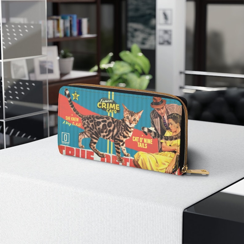 Pu&ograve; includere: Un portafoglio con cerniera blu e rosso con un'illustrazione vintage di un gatto, un uomo e una donna. Il testo sul portafoglio recita "Famous Crime Cats", "Cat O' Nine Tails" e "She Knew A Way To Kill".