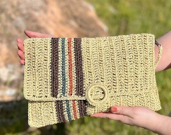 May include: A beige woven clutch with a flap closure and a circular clasp. The clutch features vertical stripes in brown, orange, and teal. The bag is held up against a blurred green background.