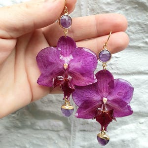 May include: A pair of handcrafted earrings featuring vibrant purple orchid petals. Each earring has a gold-colored hook, a round purple gemstone, and a heart-shaped purple gemstone pendant. The earrings are held against a white brick wall.