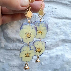 May include: Gold-colored dangle earrings featuring pressed yellow and purple pansy flowers encased in clear resin. The earrings have a sunburst design at the top and small, gold-capped clear beads at the bottom.