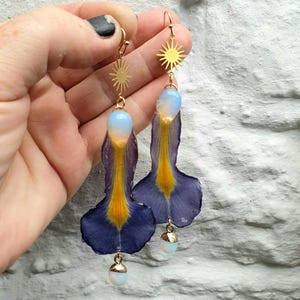Può includere: Orecchini pendenti con un ciondolo a forma di stella solare, un petalo blu e giallo e una perla bianca a forma di goccia. Gli orecchini sono sospesi a ganci color oro.