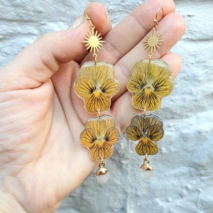 Peut inclure: Une paire de boucles d'oreilles en or avec un charme en forme d'étoile en haut et deux fleurs de pensées jaunes en résine. Les boucles d'oreilles ont une perle blanche en bas.