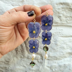 Può includere: Orecchini pendenti lunghi con viole del pensiero viola pressate, racchiuse in resina trasparente. Gli orecchini hanno ganci color oro e sono impreziositi da perle bianche e ciondoli a forma di conchiglia di abalone iridescente.