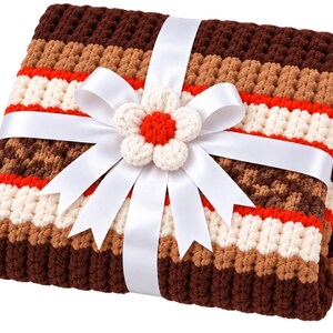 May include: A folded crochet blanket featuring horizontal stripes in dark brown, light brown, cream, and bright orange. The blanket is wrapped with a white satin ribbon tied in a bow, centered with a small, handmade cream and orange flower.