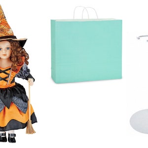 May include: A porcelain doll dressed as a witch with an orange and black dress, a witch's hat, and a broom. The doll is standing on a white doll stand. A light blue paper bag is also included.