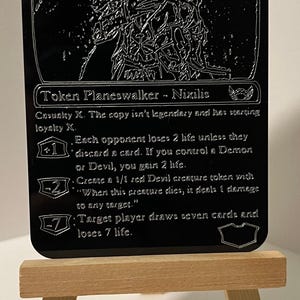 May include: Black and white image of a Magic: The Gathering card featuring Ob Nixilis, the Adversary. The card is a planeswalker with a starting loyalty of 7. The card text reads: "Casualty X. The copy isn't legendary and has starting loyalty X. +1: Each opponent loses 2 life unless they discard a card. If you control a Demon or Devil, you gain 2 life. -2: Create a 1/1 red Devil creature token with "When this creature dies, it deals 1 damage to any target." -7: Target player draws seven cards and loses 7 life."
