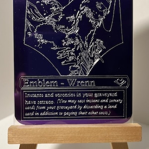 May include: A purple Magic: The Gathering card with silver foil lettering. The card is an emblem for Wrenn. The text reads "Emblem - Wrenn. Instants and sorceries in your graveyard have retrace. (You may cast instant and sorcery cards from your graveyard by discarding a land card in addition to paying their other costs.)"