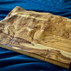 Könnte beinhalten: Rechteckiges Servierbrett aus Holz mit einem eingearbeiteten topografischen Design. Das Brett besteht aus hellem und dunklem braunem Holz mit einer detaillierten Umrisszeichnung eines Gebirges. Das Brett wird auf einer blauen Oberfläche präsentiert.