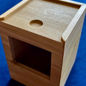 Può includere: Una piccola scatola di legno con un coperchio incernierato e un ritaglio circolare nella parte superiore. La scatola ha un'apertura rettangolare nella parte anteriore.