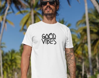 May include: A white short-sleeved t-shirt featuring the black text "GOOD VIBES" printed on the chest, paired with multi-colored shorts displaying a vibrant tropical leaf pattern in shades of pink, purple, teal, and blue.