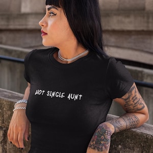 May include: A black short-sleeved t-shirt featuring the white text "HOT SINGLE AUNT" printed across the chest in a stylized, slightly distressed font. The garment has a classic crew neckline and a fitted silhouette, made of a solid black fabric.