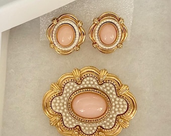 May include: A jewelry set consisting of a brooch and a pair of matching earrings. Each piece features a central oval light pink cabochon stone surrounded by a border of tiny white faux pearls, all set within a scalloped gold-tone metal frame.