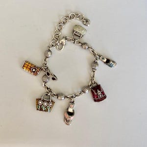 May include: Silver charm bracelet with multiple charms, including a handbag, a high heel, a purse, and a small suitcase. The charms are in various colours, including red, pink, and gold. The bracelet is on a white surface.