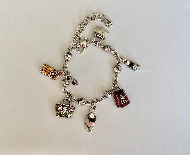 May include: Silver charm bracelet with multiple charms, including a handbag, a high heel, a purse, and a small suitcase. The charms are in various colours, including red, pink, and gold. The bracelet is on a white surface.