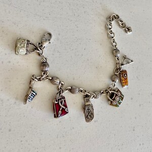 May include: Silver charm bracelet featuring multiple charms, such as a handbag, a shoe, a lipstick, and a red bag. The charms are detailed with colourful enamel and intricate designs. The bracelet is on a white surface.