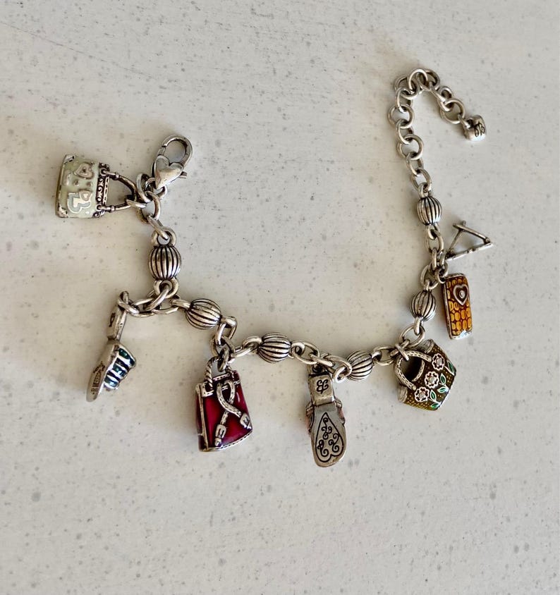 May include: Silver charm bracelet featuring multiple charms, such as a handbag, a shoe, a lipstick, and a red bag. The charms are detailed with colourful enamel and intricate designs. The bracelet is on a white surface.