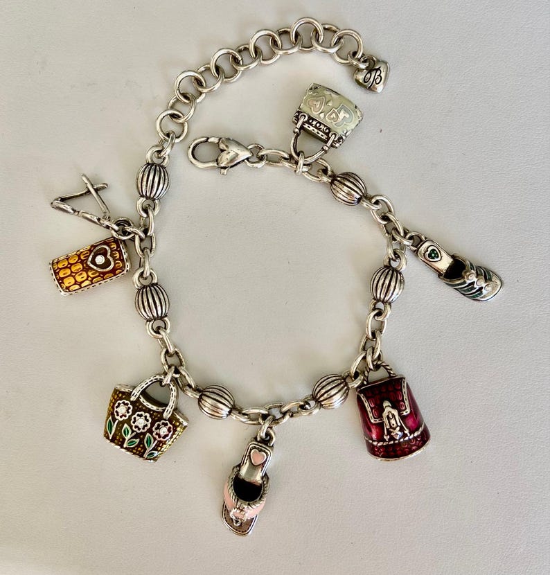 May include: Silver charm bracelet with multiple charms, such as a handbag, a shoe, and a purse. The charms are detailed with colourful enamel and intricate designs. The bracelet has a lobster clasp closure and is made of silver-toned metal.