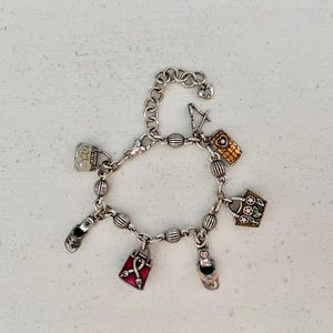 May include: Silver charm bracelet with multiple charms, such as handbags, a shoe, a high heel, and a mobile phone. The charms have colourful details and textures. The bracelet is shown on a white surface.