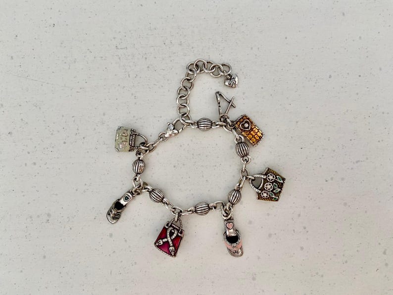 May include: Silver charm bracelet with multiple charms, such as handbags, a shoe, a high heel, and a mobile phone. The charms have colourful details and textures. The bracelet is shown on a white surface.