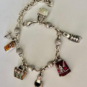 May include: Silver charm bracelet with a variety of charms, such as a handbag, shoe, and purse. The bracelet has a lobster clasp and a decorative chain. The charms are detailed with colourful enamel and rhinestones.