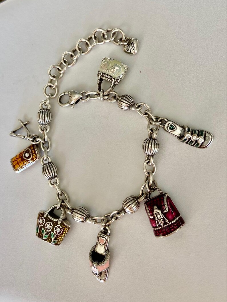 May include: Silver charm bracelet with a variety of charms, such as a handbag, shoe, and purse. The bracelet has a lobster clasp and a decorative chain. The charms are detailed with colourful enamel and rhinestones.