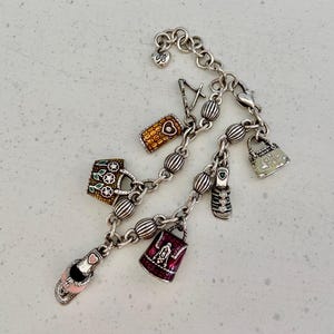 May include: Silver charm bracelet with various charms, including a handbag, a shoe, and a phone. The charms are detailed with colourful enamel and decorative elements. The bracelet is on a light-coloured surface.