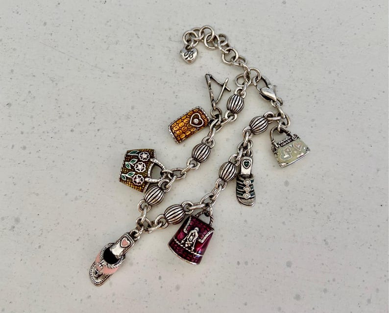 May include: Silver charm bracelet with various charms, including a handbag, a shoe, and a phone. The charms are detailed with colourful enamel and decorative elements. The bracelet is on a light-coloured surface.