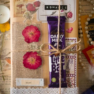 May include: A rectangular package wrapped in brown paper, decorated with pressed red flowers, vintage-style stickers, and a white lace trim. A purple Cadbury Dairy Milk Little Bar is tied to the front with twine. The text ROMANCE and HEART are visible.