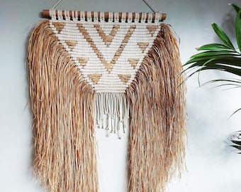 Könnte beinhalten: Ein Wandbehang aus Makramee mit einem geometrischen Muster in Beige und Weiß. Der Behang hat unten lange, braune Fransen.
