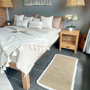 May include: A bedroom with a bed with a white and brown striped blanket, a white rug, a wooden nightstand, and a woven wall hanging with tassels.