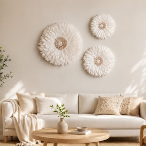 Puede incluir: Un salón de tonos neutros con un sofá blanco, cojines y una mesa de centro de madera. Tres decoraciones murales circulares con plumas blancas y centros tejidos adornan la pared. También se ven una maceta y una cesta tejida.