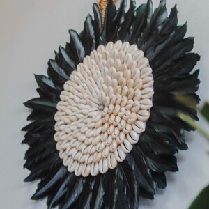 Peut inclure: Une suspension murale circulaire faite de plumes noires et de coquillages de cauris blancs. Les coquillages sont disposés en spirale.