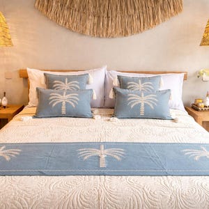Puede incluir: Una escena de dormitorio con una cama vestida con un edredón crema estampado y cojines azul claro y un camino de cama, todos adornados con un diseño de palmera blanca. Dos lámparas colgantes tejidas cuelgan sobre la cama y dos mesitas de noche de madera flanquean la cama.
