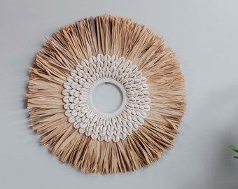 May include: A round wall hanging with a natural, woven raffia design. The center features a ring of white seashells, surrounded by a larger circle of raffia. The raffia is a light tan color, and the seashells are white.