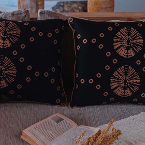 May include: Two black throw pillows with a geometric pattern of brown circles and small squares. The pillows are on a couch with a book and a dried flower arrangement.