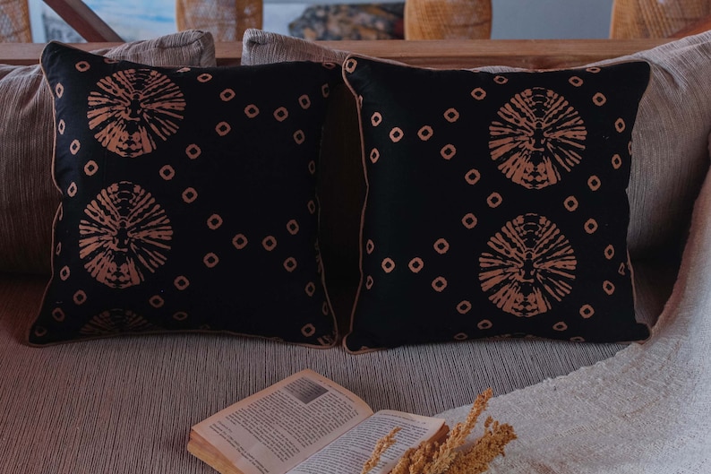 May include: Two black throw pillows with a geometric pattern of brown circles and small squares. The pillows are on a couch with a book and a dried flower arrangement.