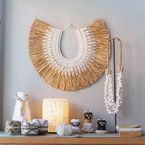 Può includere: Un mobile in legno con due ante e uno scaffale. L'armadio ha un design in rattan intrecciato sulle porte. Sullo scaffale ci sono diversi oggetti decorativi, tra cui un'appendice da parete in paglia e conchiglie, una collana di conchiglie bianche, una lampada di conchiglie bianche e tre piccole figure intrecciate.