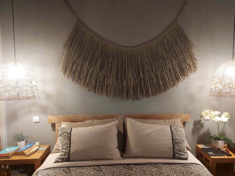 May include: A bedroom with a bed with two pillows and a woven wall hanging above the headboard. The bed has a wooden frame and a patterned bedspread. There are two bedside tables with lamps and plants.