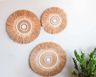May include: Three round wall decorations with a natural, fringed design. Each piece features a central woven circle adorned with white beads and a shell-like centerpiece. The neutral tones and textures create a bohemian aesthetic.