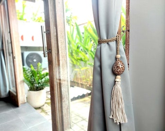 May include: A decorative curtain tieback featuring a natural seashell with a brown spotted pattern, wooden beads, and a cream-colored tassel. The tieback is tied around a gray curtain, adding a touch of coastal decor.