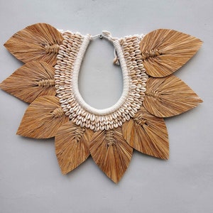 May include: A handcrafted statement necklace featuring a central collar of small, white seashells. The necklace is surrounded by large, stylized leaf shapes made of natural, light brown fibers. The design is set against a neutral gray background.