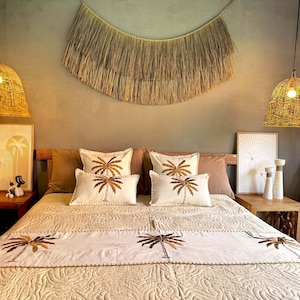 Puede incluir: Una cama blanca con una colcha de color crema con palmeras bordadas. La cama tiene una cabecera marrón y está decorada con almohadas blancas y marrones. La habitación tiene un tema bohemio con lámparas de tejido y una pared de tejido.