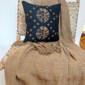 May include: A brown throw blanket with fringe edges draped over a chair with a black and tan patterned pillow. The chair has a tan and brown patterned fabric upholstery.
