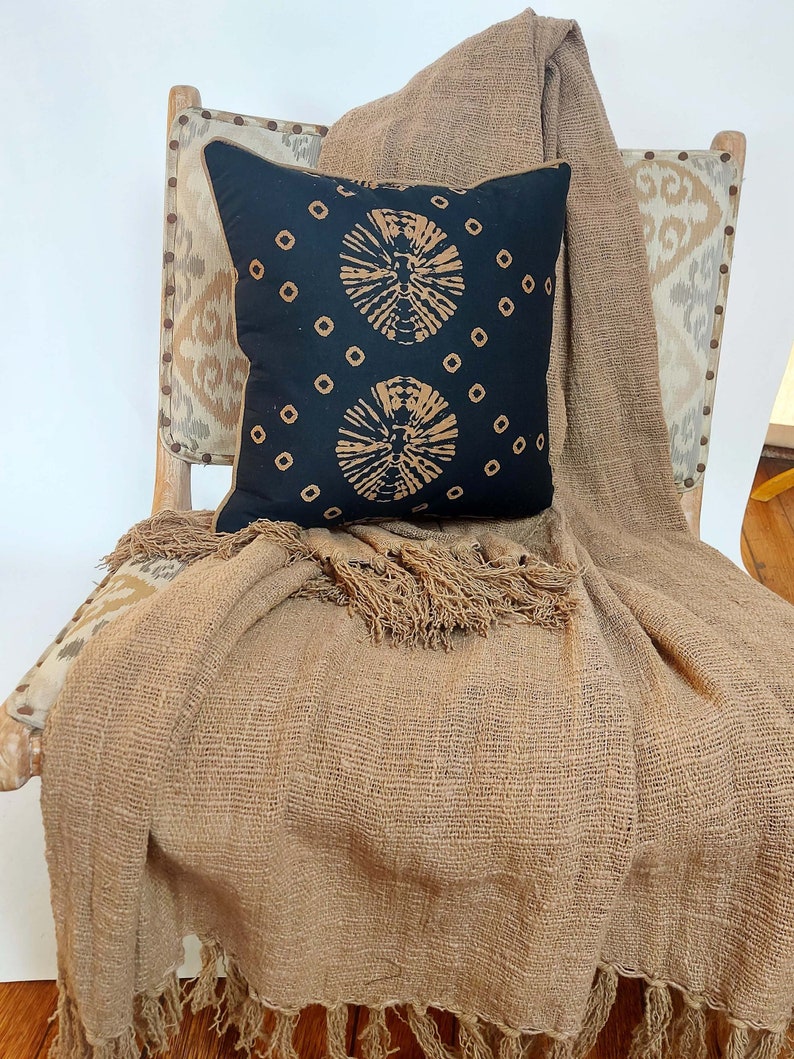 May include: A brown throw blanket with fringe edges draped over a chair with a black and tan patterned pillow. The chair has a tan and brown patterned fabric upholstery.