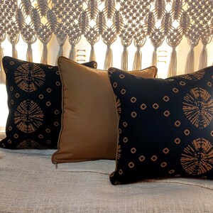 May include: Three decorative throw pillows with a black background and a brown geometric pattern. The pillows are arranged on a beige textured surface.